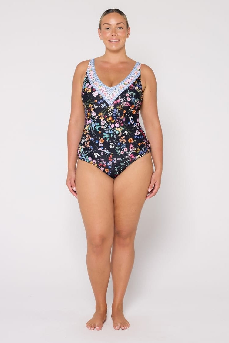 A woman modeling a one-piece swimsuit with a black background and a colorful floral print, featuring a light blue and white patterned V-neckline trim, against a plain light gray studio background.