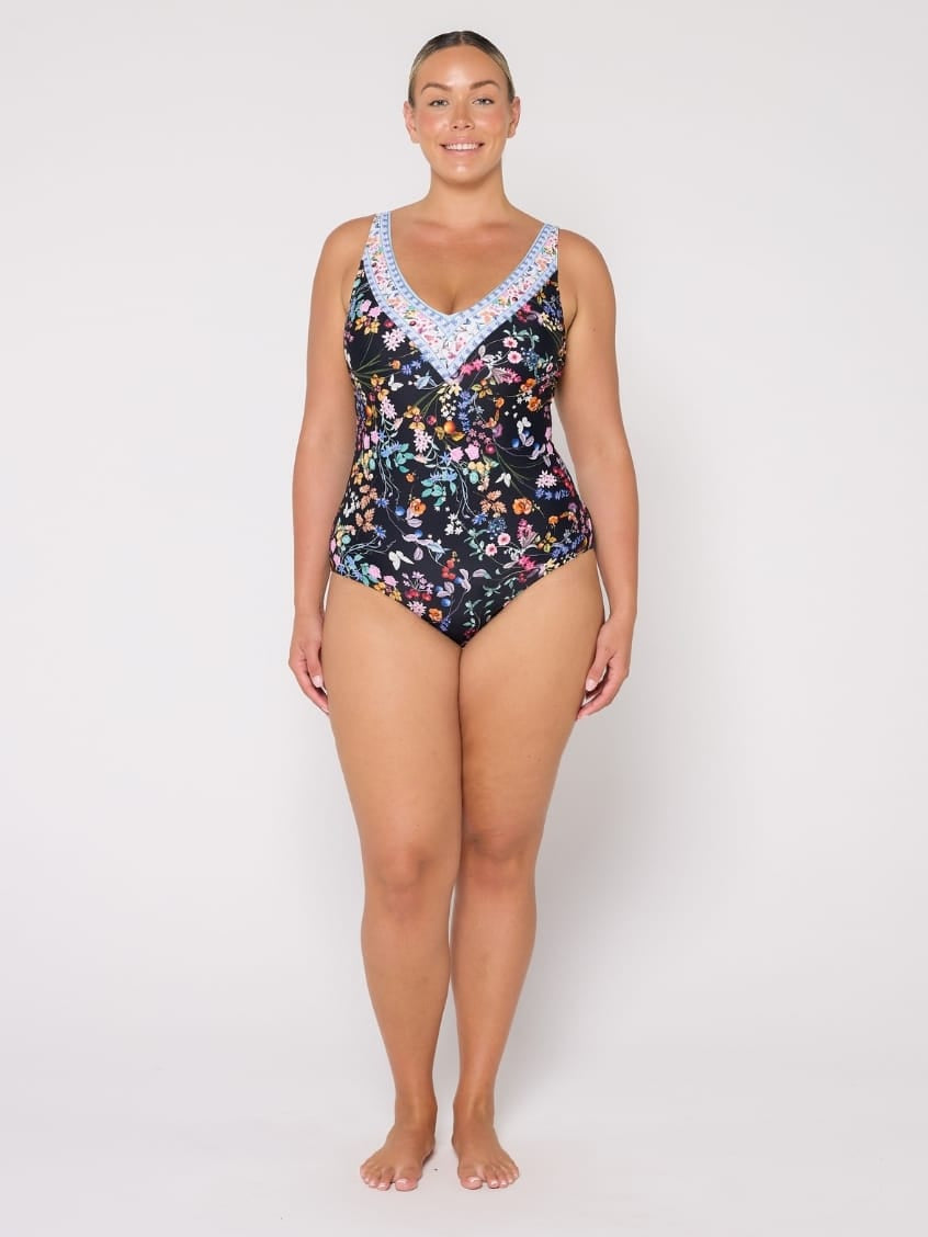 A woman modeling a one-piece swimsuit with a black background and a colorful floral print, featuring a light blue and white patterned V-neckline trim, against a plain light gray studio background.