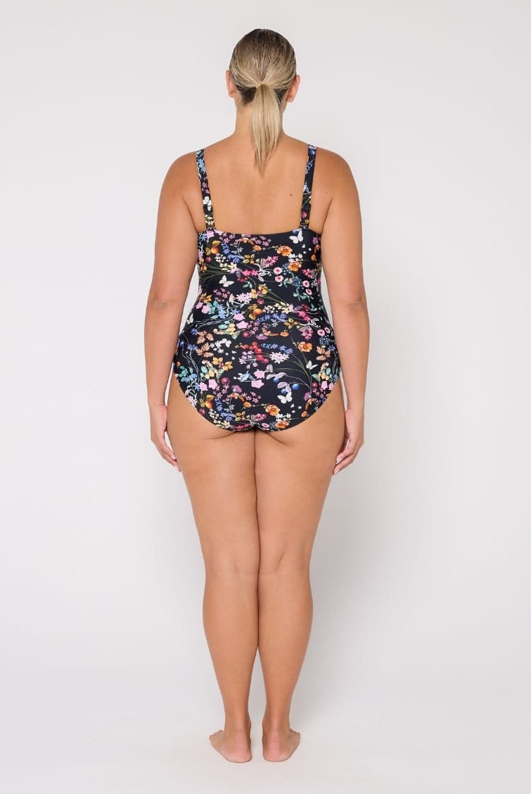 A woman viewed from the back wearing a one-piece swimsuit with a dark floral print, standing against a plain light gray studio background.