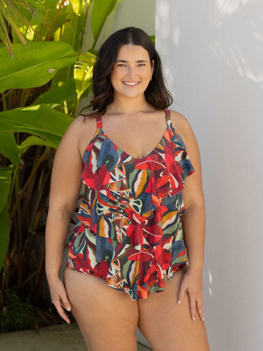 A woman modeling a colorful floral print ruffled one-piece swimsuit, standing outdoors against a white wall with tropical green leaves on the left.