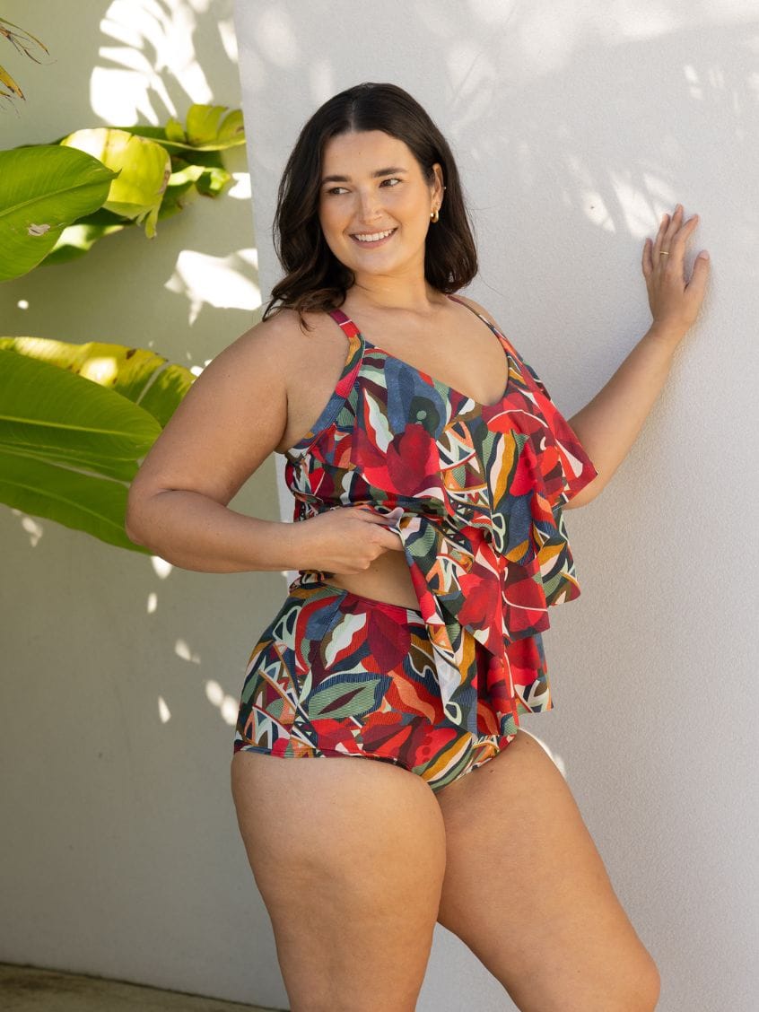 A woman modeling a colorful floral print halter swimsuit with ruffle details, posing against a white stucco wall with tropical leaves on the left.
