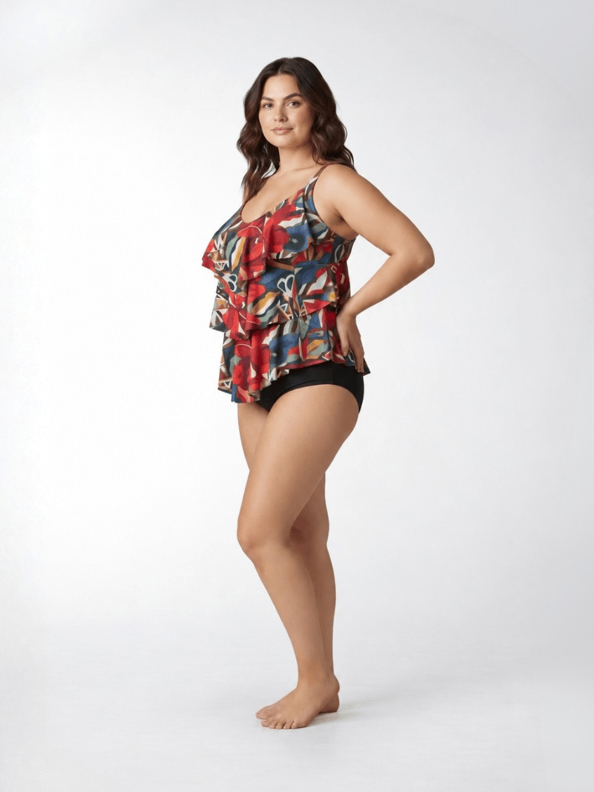 A woman modeling a sleeveless ruffled tankini top with a colorful abstract print in red, blue, yellow, and black, paired with black bikini bottoms, against a plain white studio background.