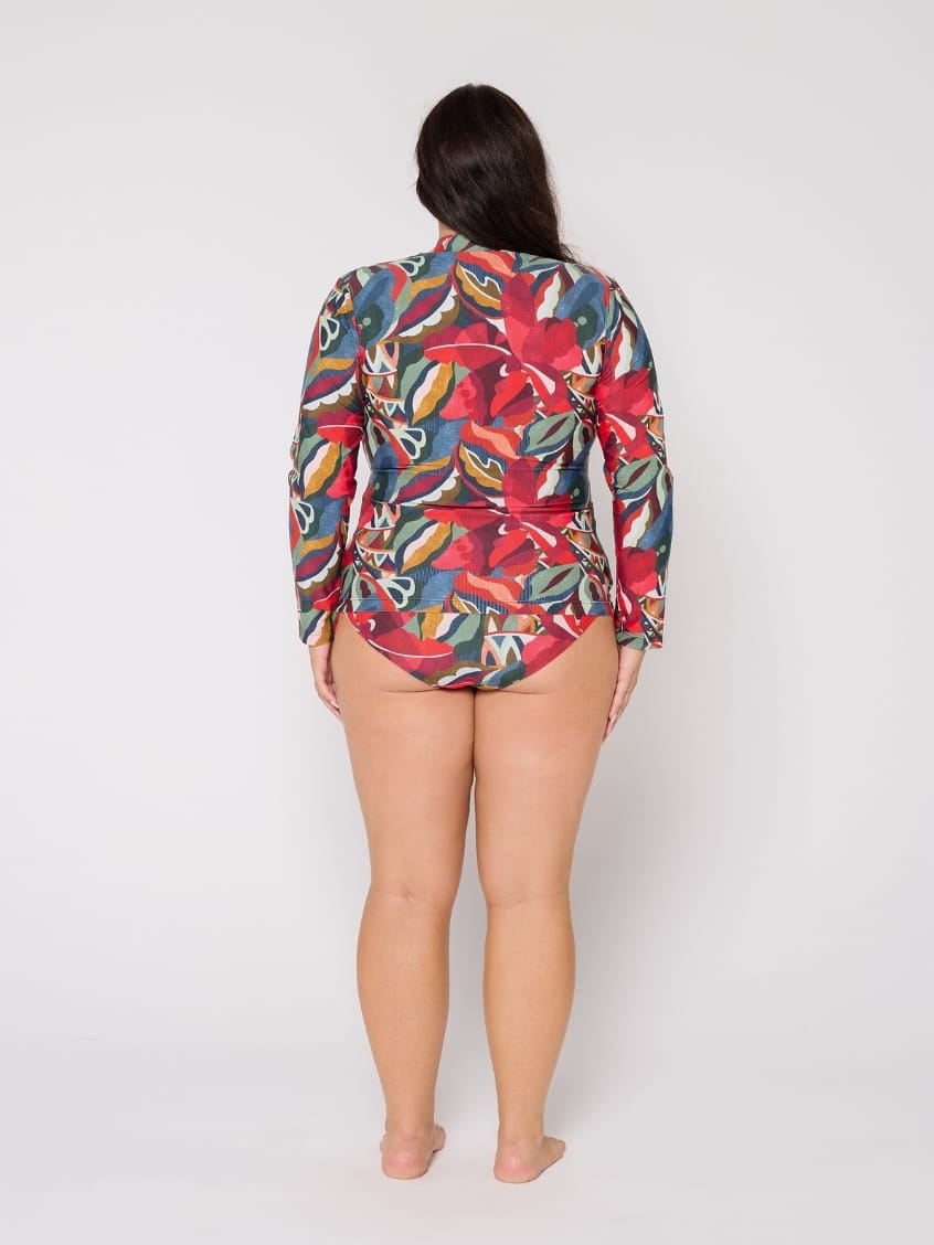 A woman viewed from behind wearing a long-sleeve, high-neck bikini top with a vibrant tropical floral print in red, blue, green, and yellow, paired with matching bikini bottoms, against a plain light gray background.