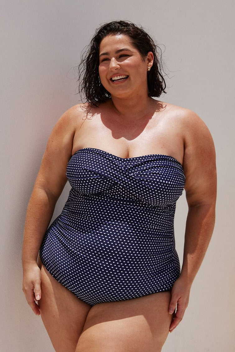 brunette model wearing a strapless one piece bathing suit with twist detail in between the breast area