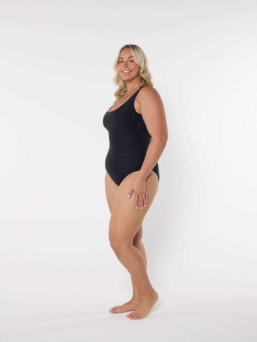 Women wearing a black one piece swimsuit standing in front of a white wall