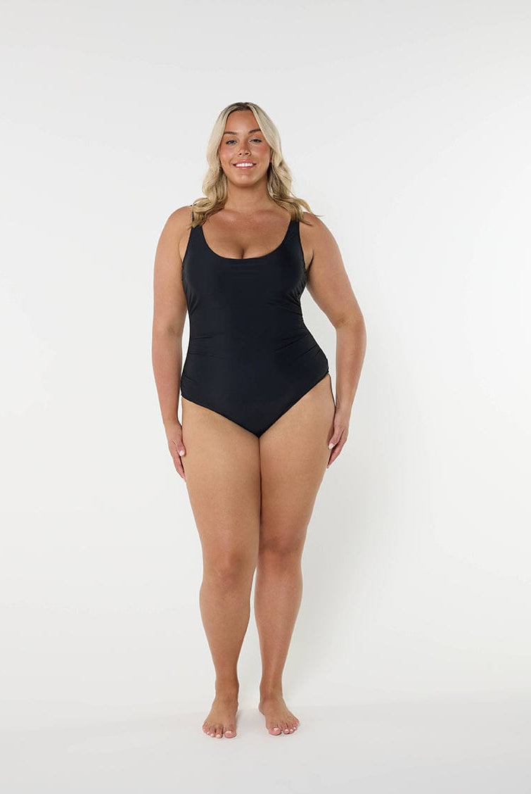 Women wearing a black one piece swimsuit standing in front of a white wall