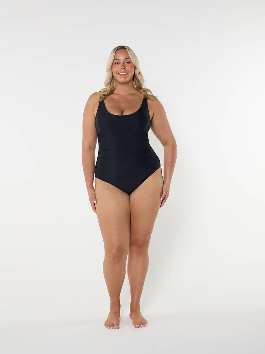 Women wearing a black one piece swimsuit standing in front of a white wall