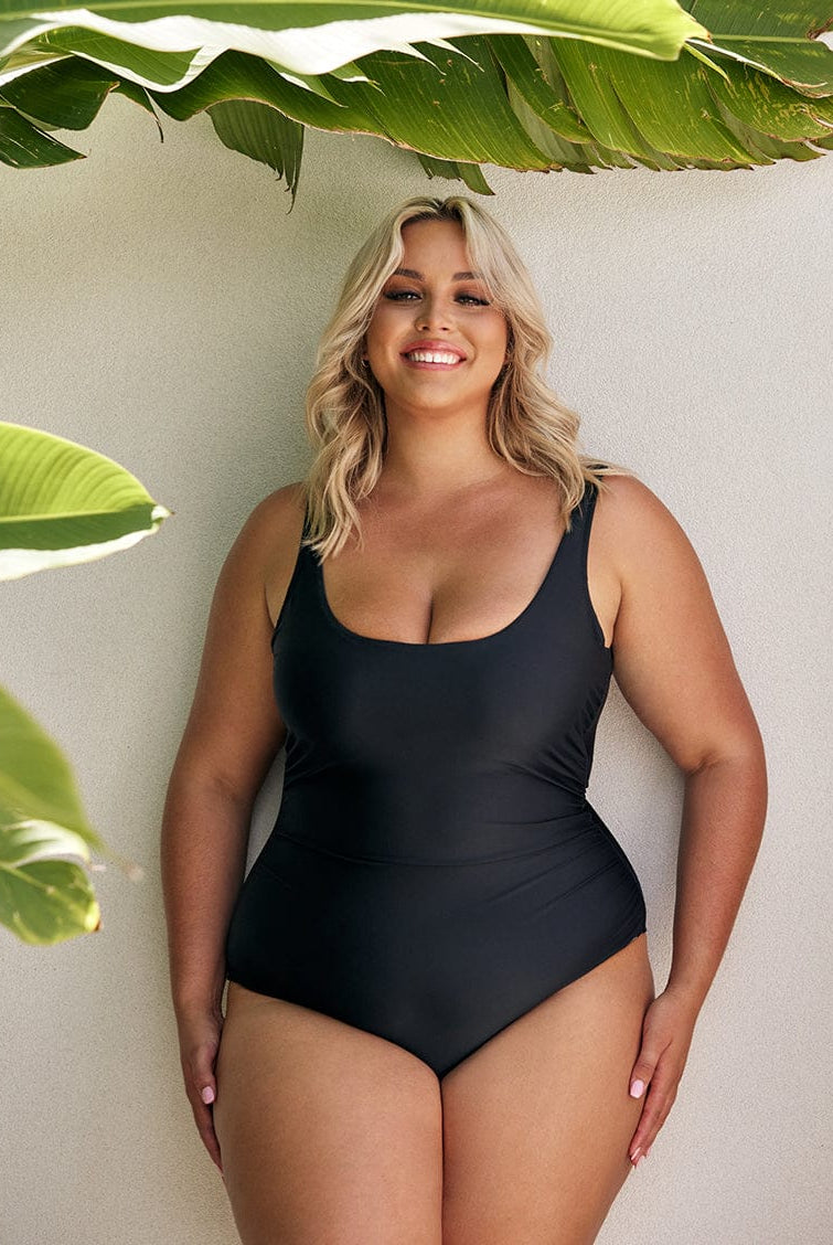 Women wearing a black one piece swimsuit standing in front of a white wall