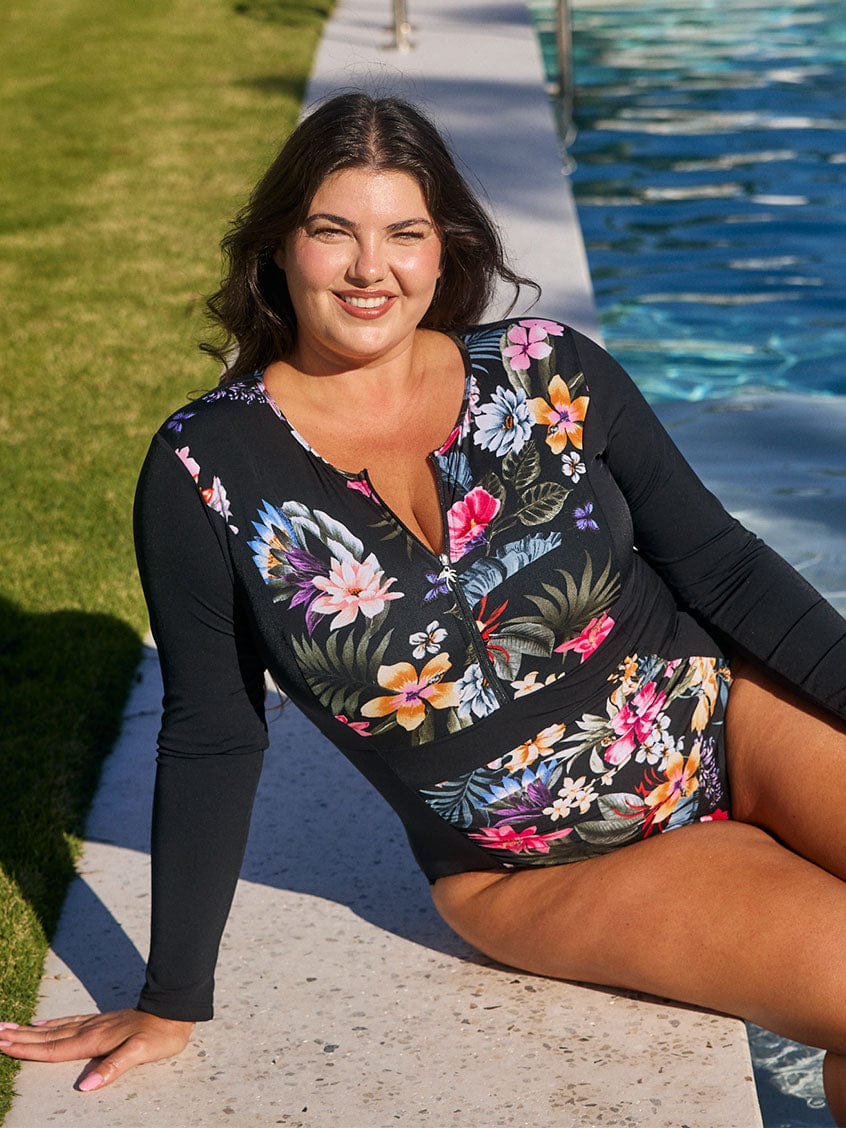 Brunette model wears zip front chlorine resistant black floral one piece with long sleeves