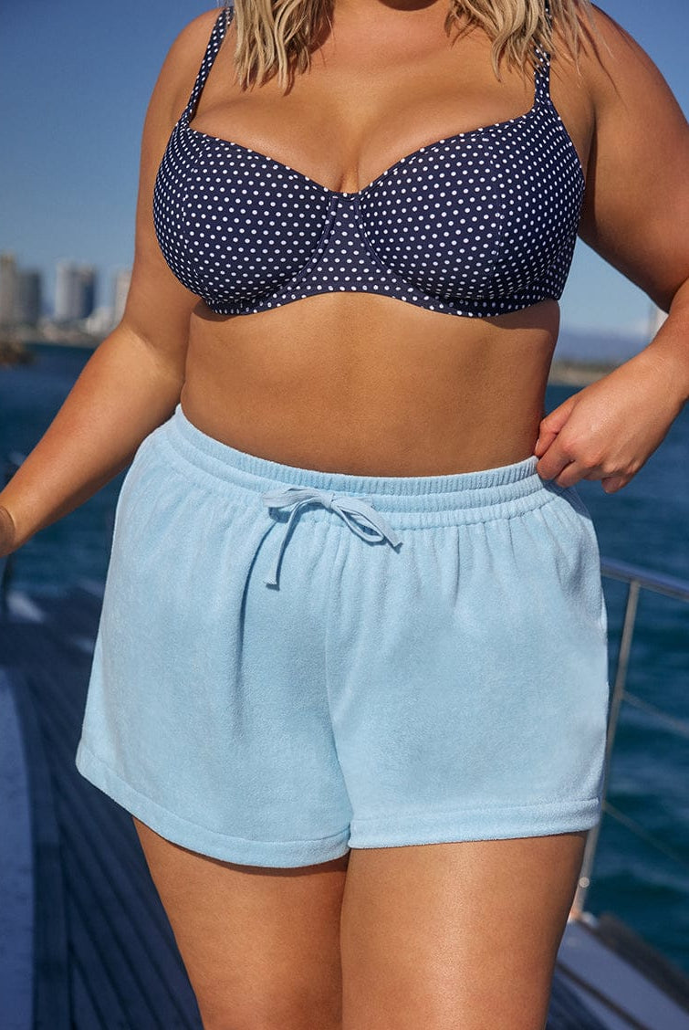 Person wearing a navy polka dot bikini top and light blue shorts on a boat with water and cityscape in the background.