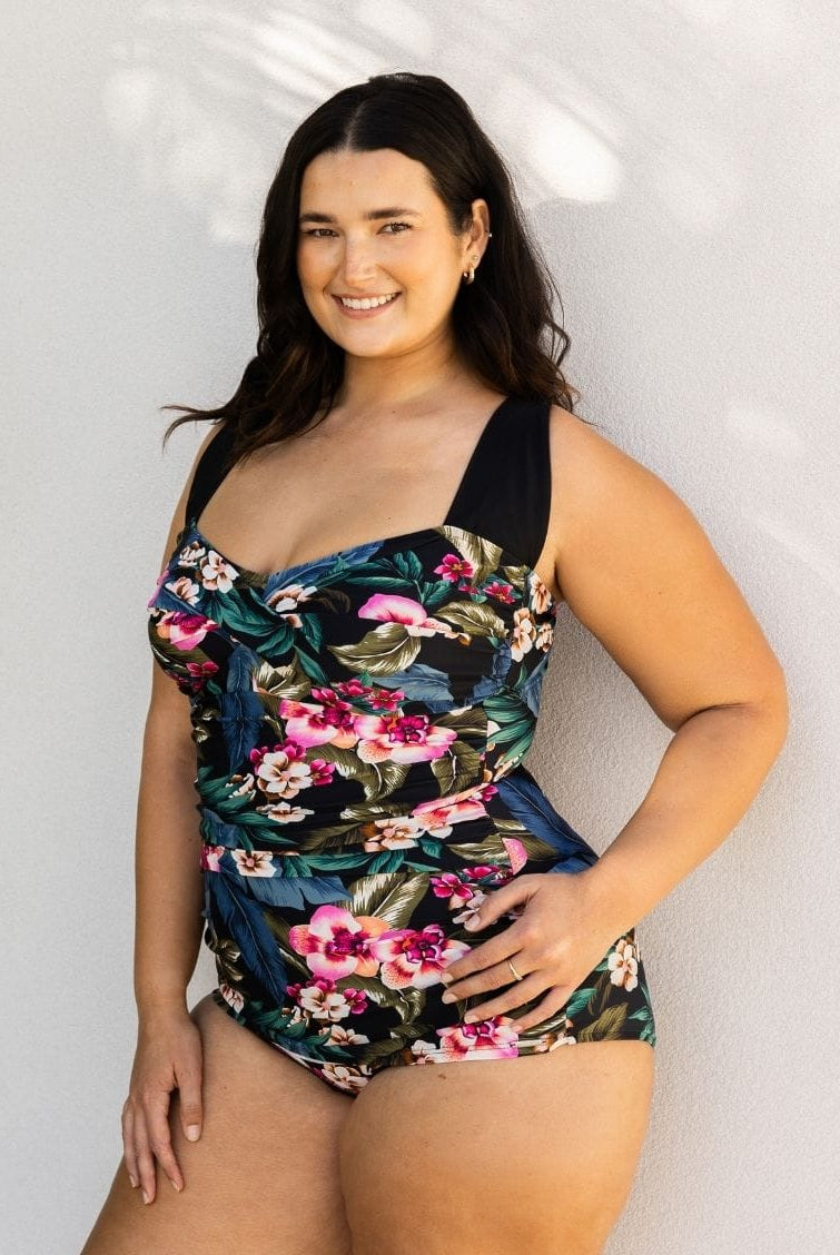 A woman modeling a one-piece swimsuit with a dark navy and black base featuring a vibrant pink, red, and green tropical floral print, posed against a light gray textured wall.