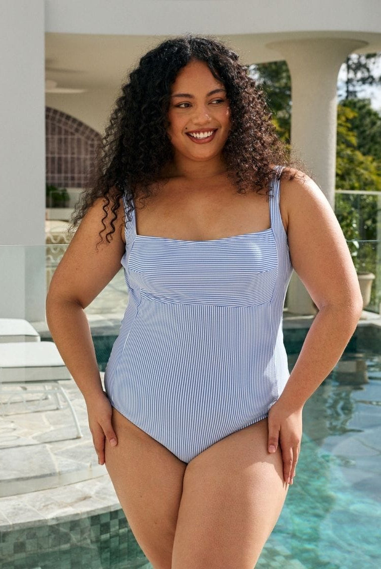 A woman modeling a light blue and white vertically striped one-piece swimsuit in front of a swimming pool and modern house patio.