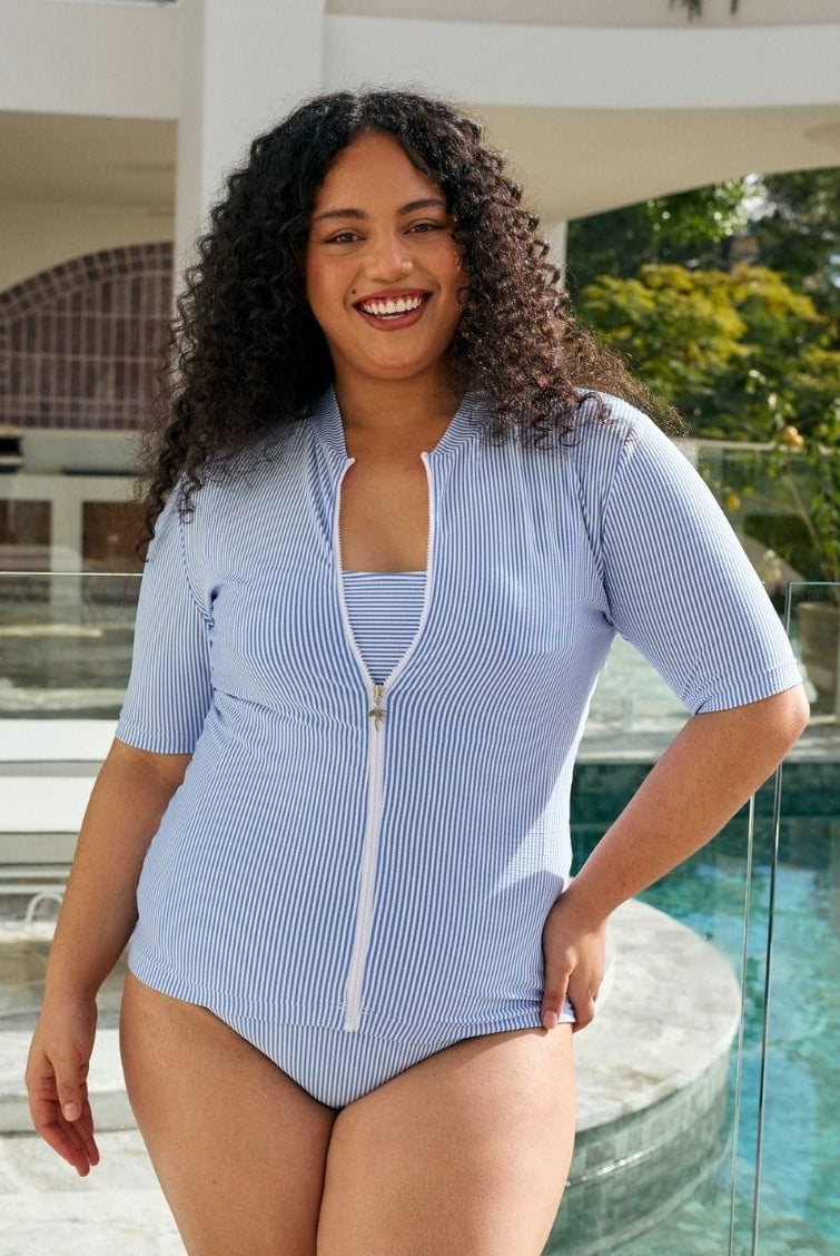A woman with curly dark hair wearing a light blue and white vertically striped one-piece swimsuit with a front zipper, standing by a swimming pool.