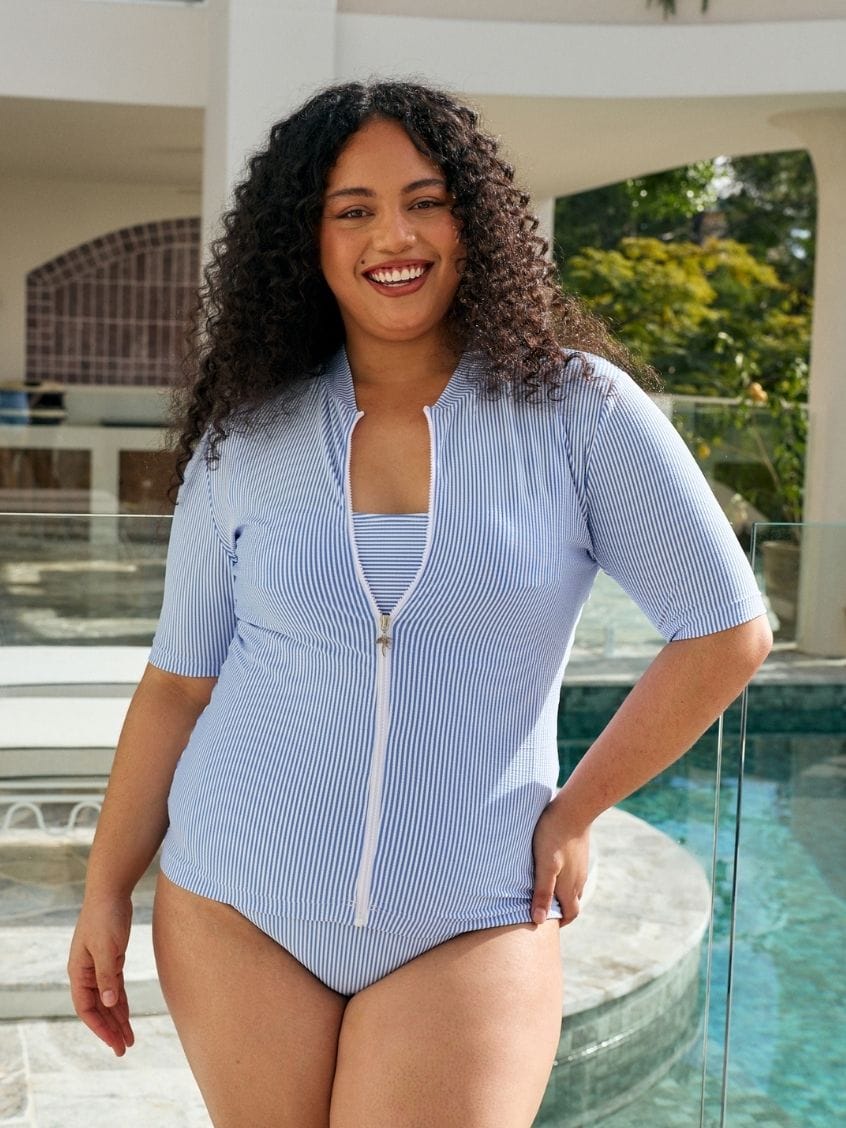 A woman with curly dark hair wearing a light blue and white vertically striped one-piece swimsuit with a front zipper, standing by a swimming pool.