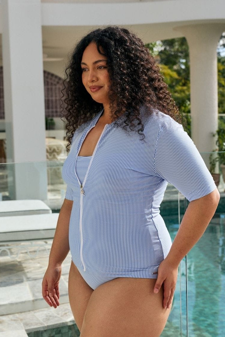 A woman modeling a light blue and white vertically striped zip-up one-piece swimsuit with short sleeves, standing by a swimming pool.