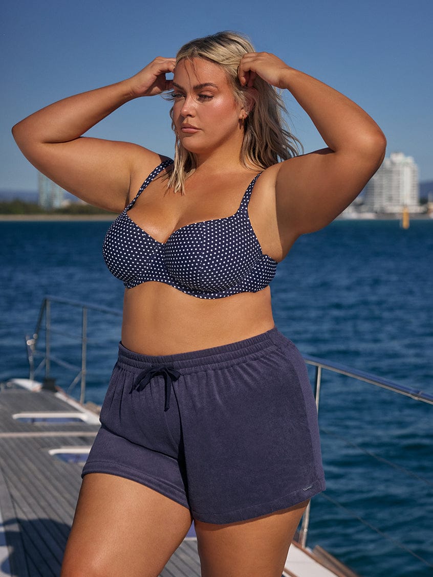 Woman in a navy polka dot bikini top and shorts standing on a boat with water and skyline in the background.