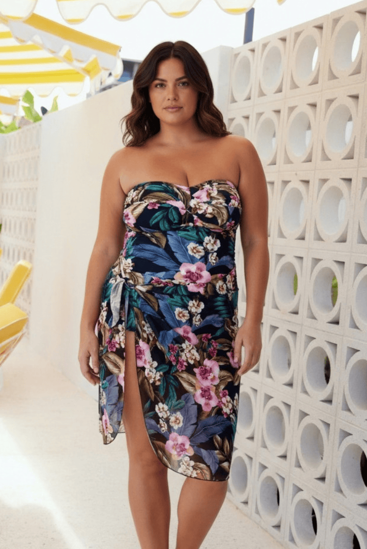 A woman models a strapless floral tube dress with a sweetheart neckline and a high side slit, set against a modern white block wall with circular cutouts