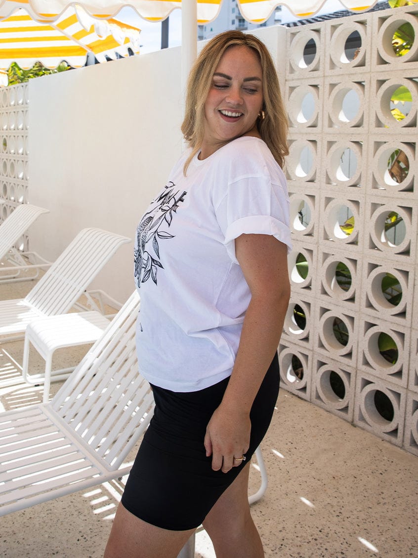 Woman wearing a white t-shirt with a graphic design and black shorts standing in front of a decorative wall.