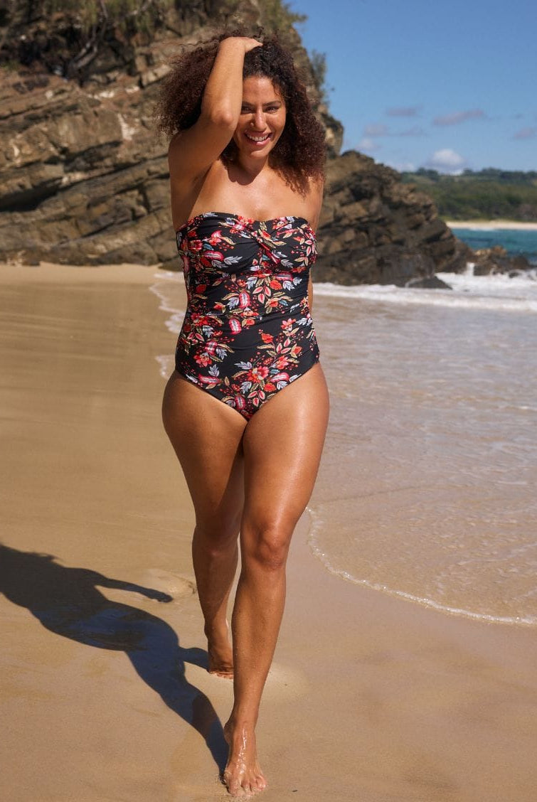 A woman with curly brown hair stands in shallow ocean water on a beach, wearing a strapless one-piece swimsuit with a black base and a colorful floral print in red, pink, and orange.