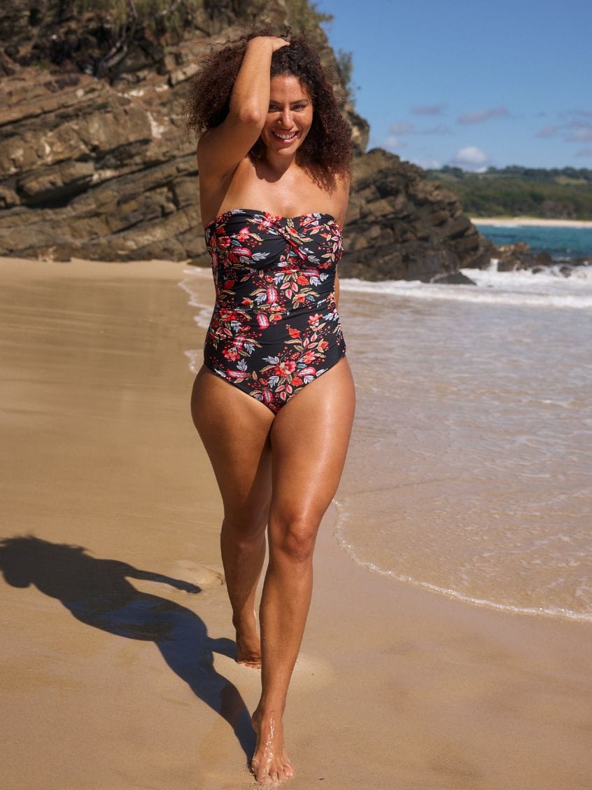A woman with curly brown hair stands in shallow ocean water on a beach, wearing a strapless one-piece swimsuit with a black base and a colorful floral print in red, pink, and orange.