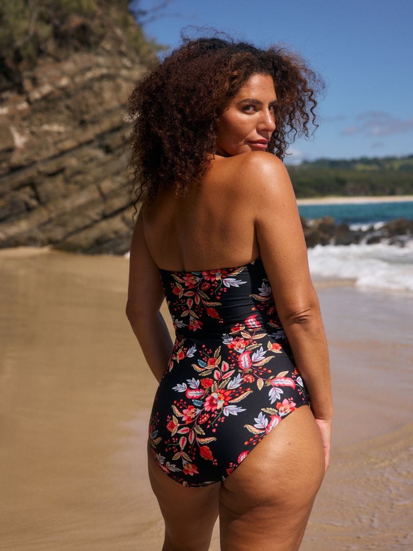A woman with curly brown hair stands on a sandy beach wearing a black strapless one-piece swimsuit with a vibrant red, pink, and white floral pattern. The background features a rocky cliff, blue ocean waves, and a clear blue sky.