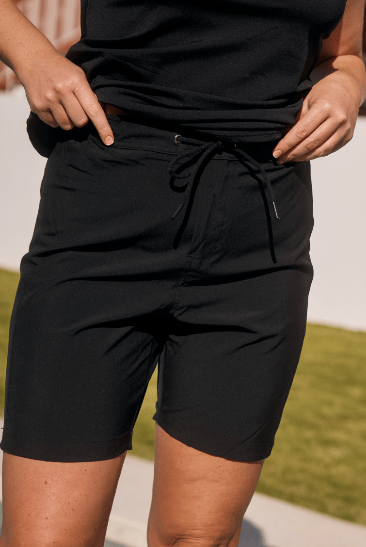 A person wearing black drawstring shorts and a black sleeveless top, standing outdoors with grass and a light wall in the background.