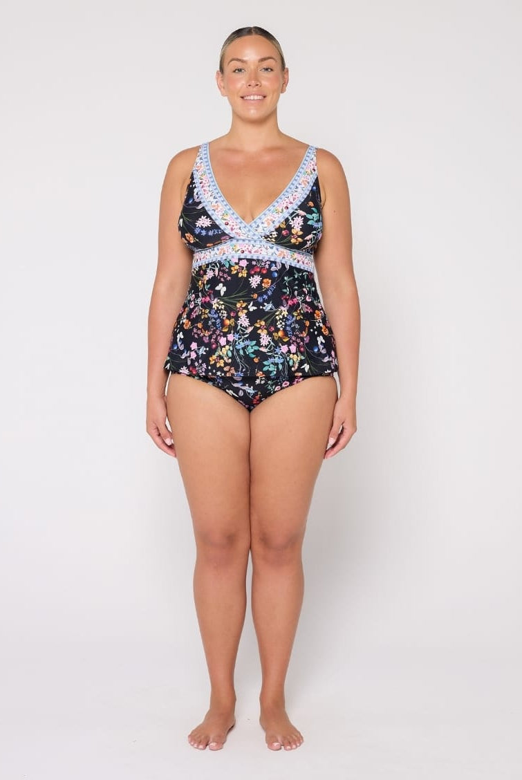 A woman modeling a one-piece swimsuit with a dark floral print and light blue crochet trim on a plain light gray studio background.
