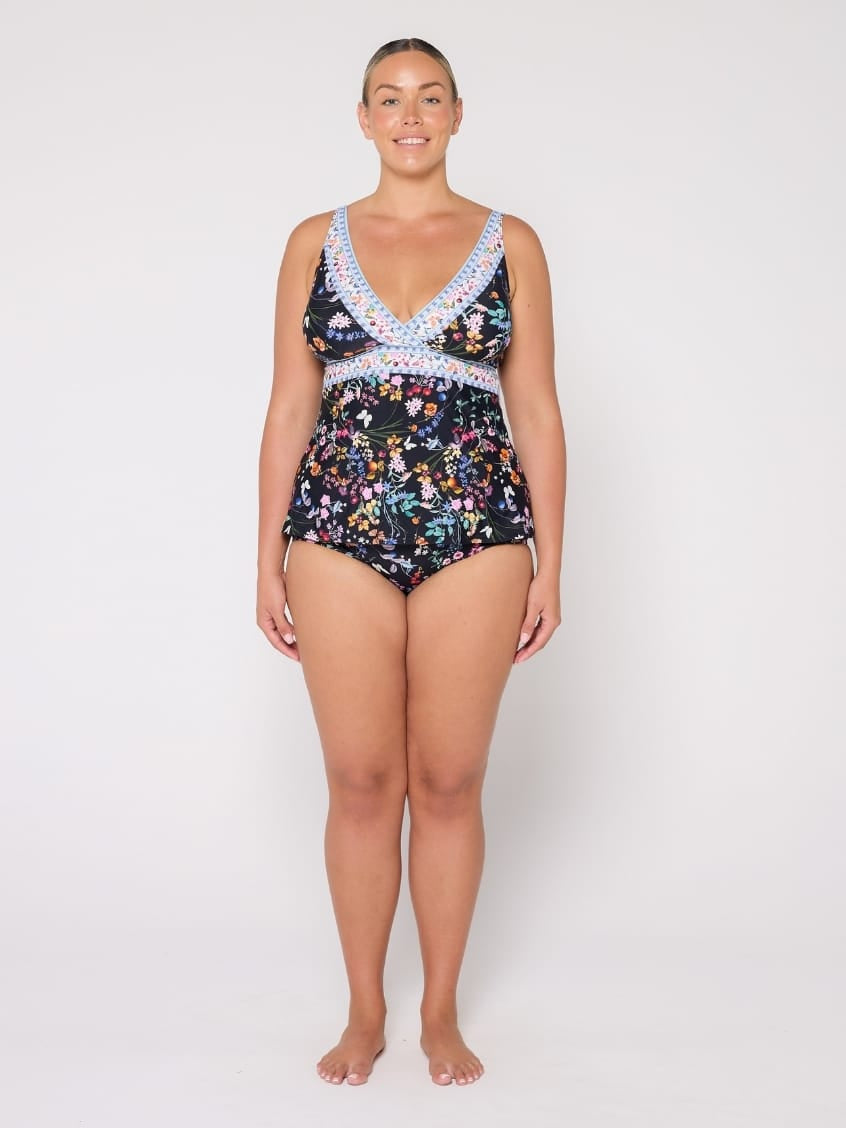 A woman modeling a one-piece swimsuit with a dark floral print and light blue crochet trim on a plain light gray studio background.