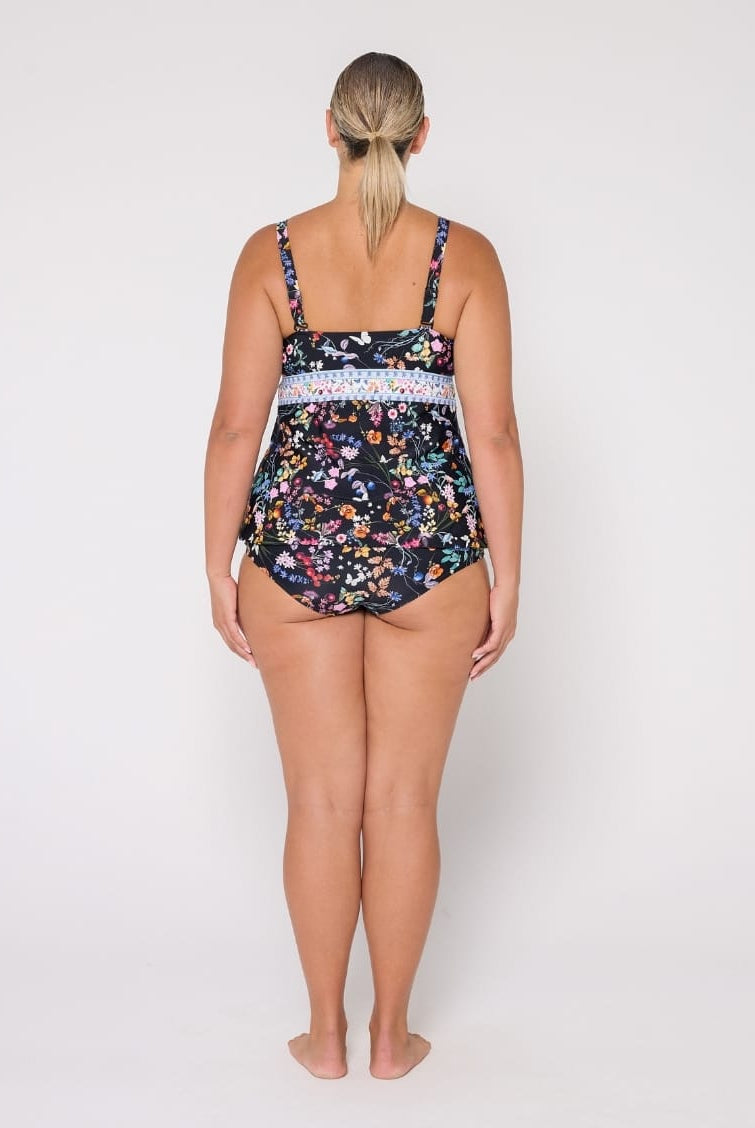 A woman stands with her back to the camera wearing a one-piece swimsuit with a dark navy floral print and a light blue patterned waistband, photographed against a plain white studio background.