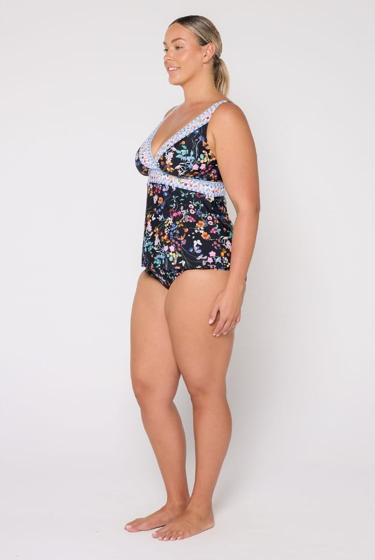 A woman modeling a one-piece swimsuit with a dark navy floral print and light blue lace trim, standing against a plain white studio background.
