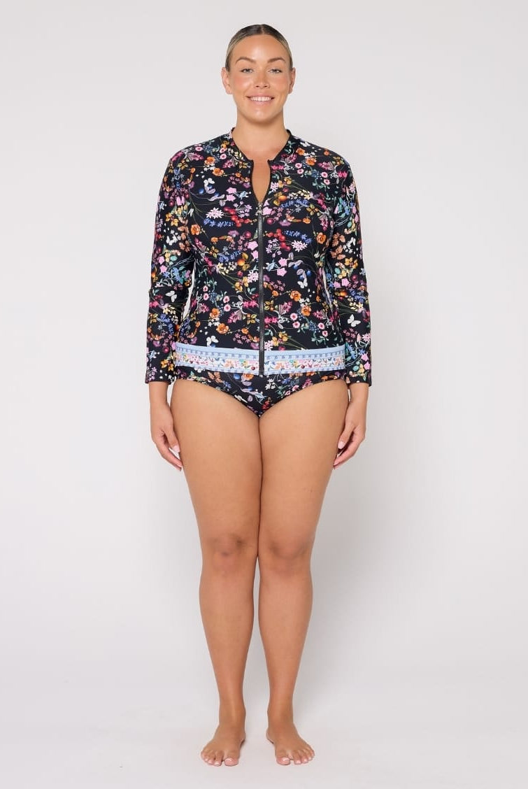 A woman modeling a two-piece swimsuit set consisting of a long-sleeve floral-print zip-front top and matching bikini bottoms, photographed against a plain light gray studio background.