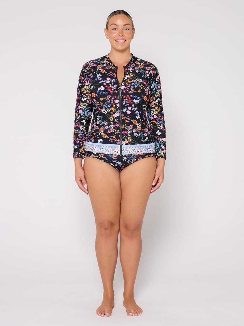 A woman modeling a two-piece swimsuit set consisting of a long-sleeve floral-print zip-front top and matching bikini bottoms, photographed against a plain light gray studio background.
