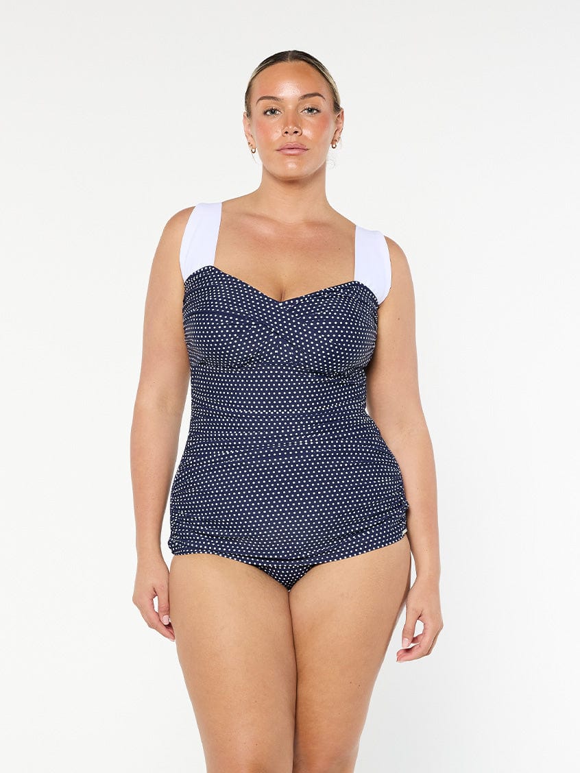 Woman wearing a navy polka dot swimsuit with white straps on a white background