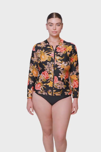 Video of brunette model wearing a long sleeve rash vest with zip front closure in a black based floral print and orange, red and yellow tones