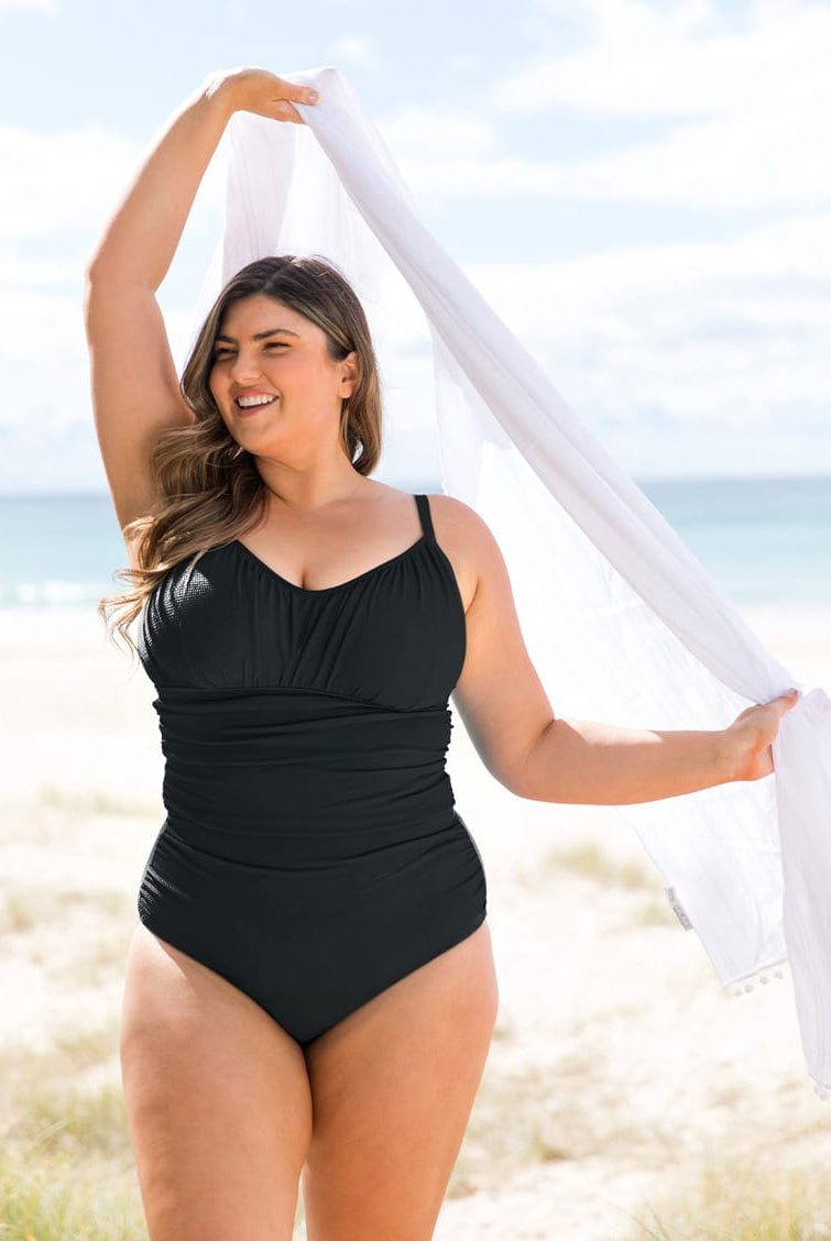 A woman stands on a sandy beach wearing a black one-piece ruched swimsuit and holding a white scarf overhead.