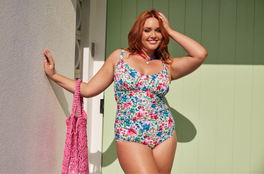 Woman with red hair wears bright coloured floral swimsuit, holds a pink woven beach bag. 