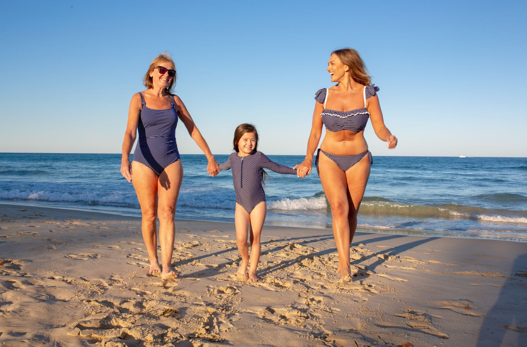 2 women and a girl walk hand in hand on the beach. They are wearing different swimsuits in a matching navy and white dots print. 