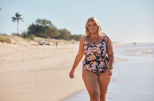 Woman with blonde hair walks along the beach wearing black, pink and orange floral swimsuit