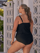 A woman wearing a black one-piece swimsuit, photographed from behind and side, standing in front of a white geometric wall with circular cutouts.