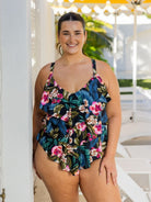 A woman modeling a one-piece tropical floral swimsuit with a ruffled hem, standing in a bright, sunny outdoor setting with white lounge chairs and a yellow umbrella in the background.