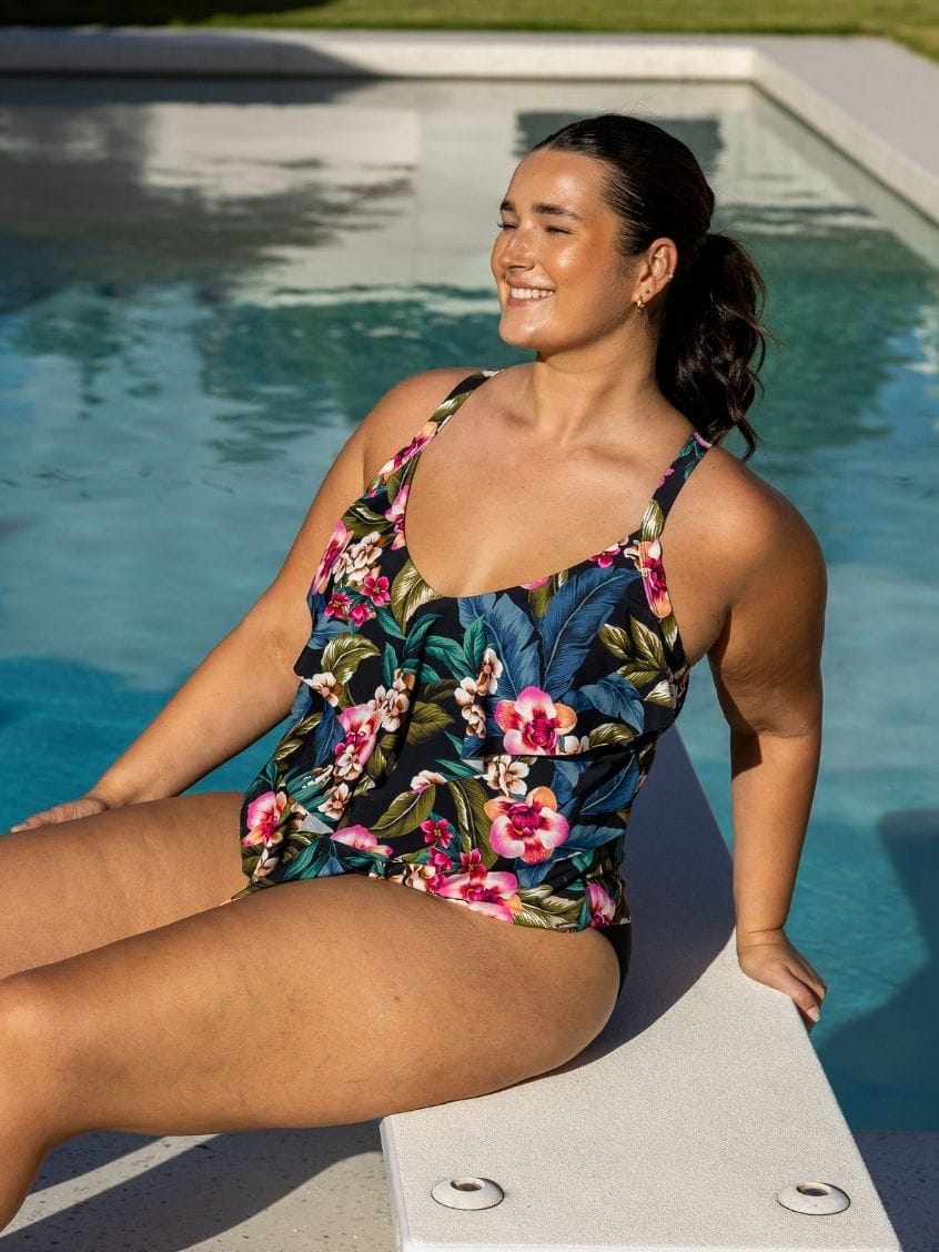 A woman wearing a floral one-piece swimsuit sits on the edge of a swimming pool outdoors.
