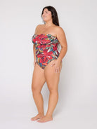 A woman modeling a strapless one-piece swimsuit with a vibrant tropical floral print in red, green, yellow, and blue, standing against a plain light gray studio background.