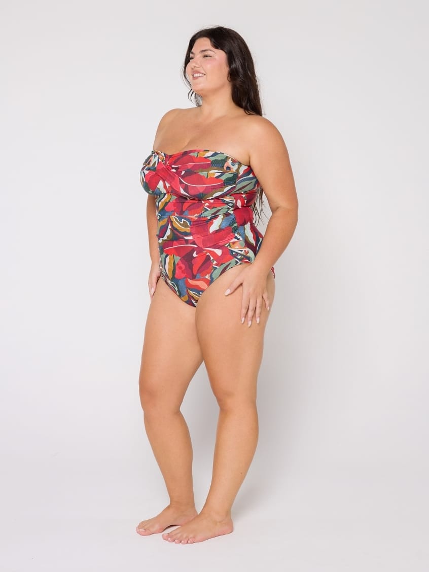 A woman modeling a strapless one-piece swimsuit with a vibrant tropical floral print in red, green, yellow, and blue, standing against a plain light gray studio background.