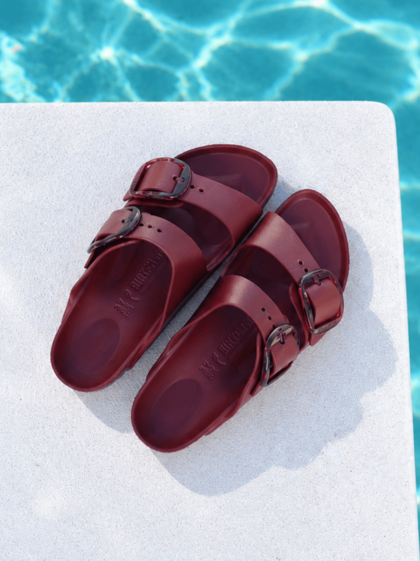A pair of burgundy double-strap slide sandals with dark metal buckles, displayed on a white stone surface with a swimming pool background.