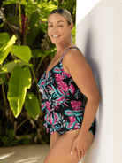 Woman wearing a tropical print swimsuit standing against a white wall with greenery in the background