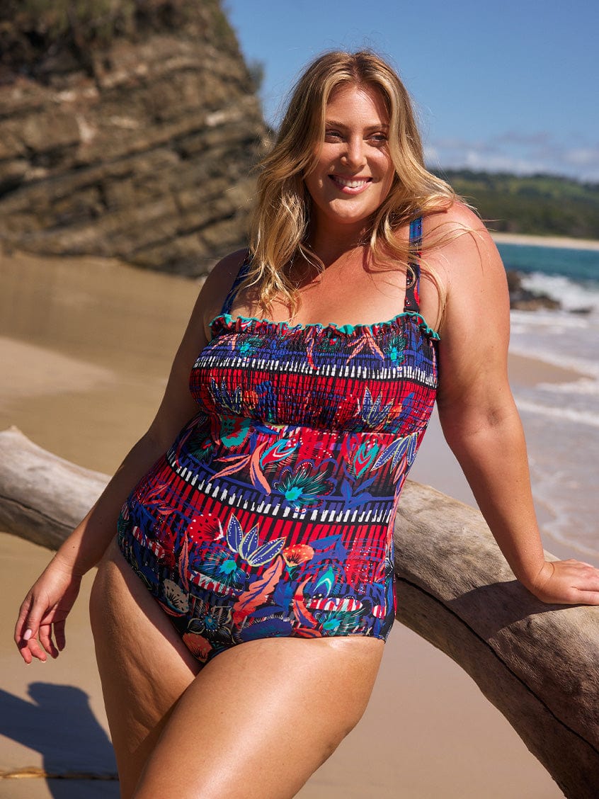 Blonde model on beach wearing a multi coloured pattern shirred one piece bandeau with straps for curve women