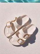 Pair of off white sandals with gold buckles on a diving board.