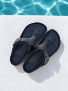 A pair of black rubber thong sandals with adjustable buckle straps, displayed on a light concrete surface with a swimming pool background.