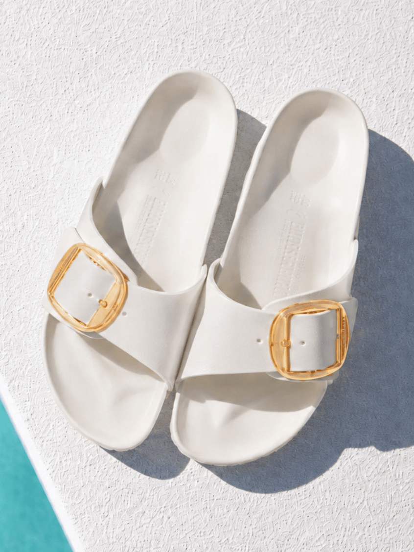 A pair of off-white slide sandals with gold square buckle details, photographed from a top-down angle on a textured white surface with a turquoise edge visible in the bottom-left corner.