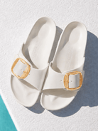 A pair of off-white slide sandals with gold square buckle details, photographed from a top-down angle on a textured white surface with a turquoise edge visible in the bottom-left corner.
